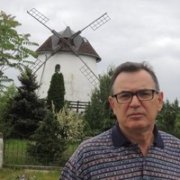 Man wearing glasses and a striped shirt stands in front of a windmill sculpture and a statue in a garden.