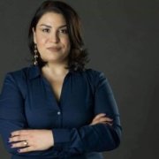 Portrait of a woman with shoulder-length dark hair, wearing a navy blue blouse and drop earrings, standing with arms crossed against a dark studio background.