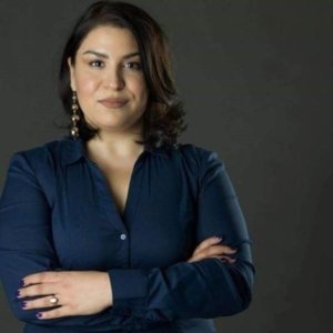 Portrait of a woman with shoulder-length dark hair, wearing a navy blue blouse and drop earrings, standing with arms crossed against a dark studio background.