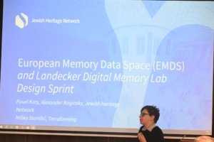 Blue conference slide with the 'Jewish Heritage Network' logo and the title 'European Memory Data Space (EMDS)'; a person stands at the right foreground.