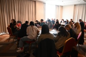 Group of people seated around tables in a bright conference room, reading and taking notes during a meeting or workshop.