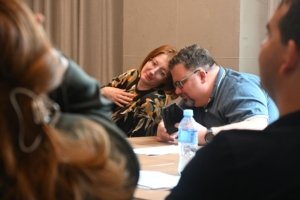 Two people lean toward each other to look at a smartphone on a table during a meeting, with a water bottle in the foreground and others blurred in the background.