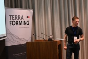 Male presenter in a black shirt standing at a wooden podium on a stage, with a Terra Forming banner on the left.