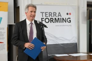 Man in a suit speaks at a microphone, holding a blue folder, with a large 'TERRA FORMING' banner behind him.