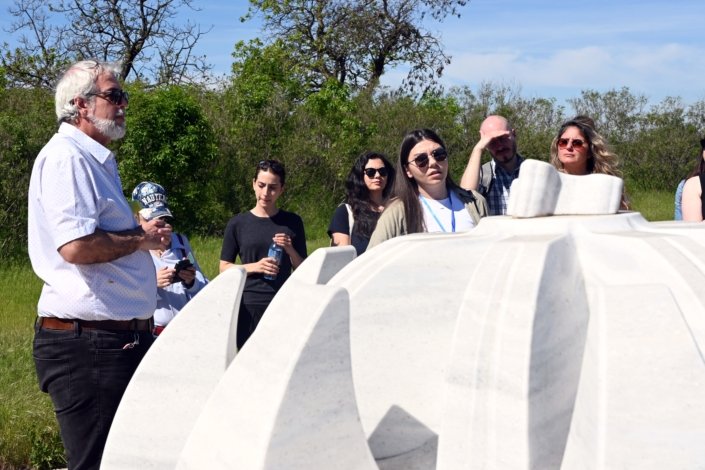 Group of adults outdoors in a grassy area, standing beside large white curved concrete forms and listening to a speaker.