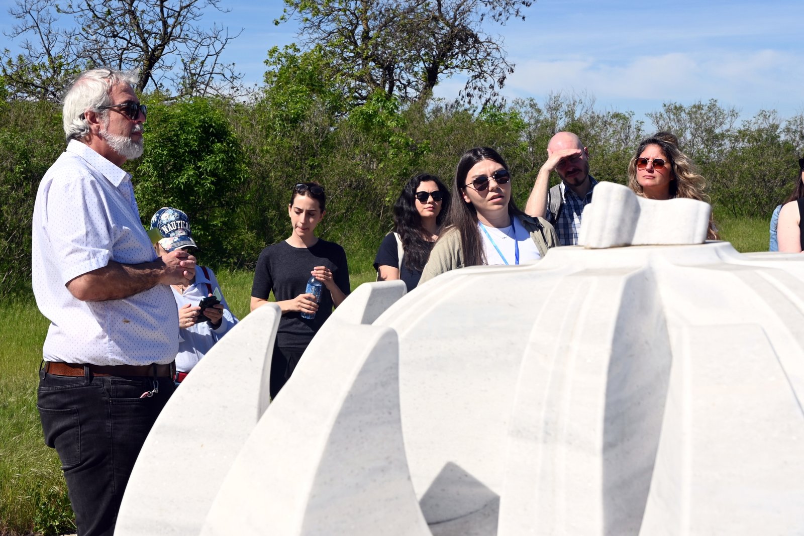 Group of adults outdoors in a grassy area, standing beside large white curved concrete forms and listening to a speaker.