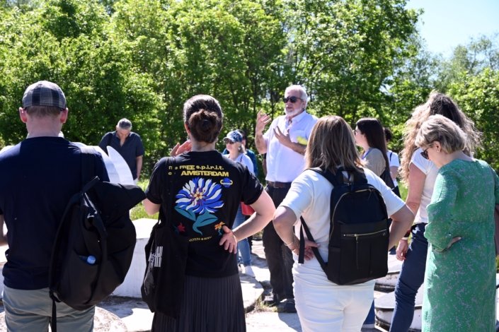 Group of people standing in a circle outdoors, listening to a man in sunglasses who is speaking to them in a sunny park.