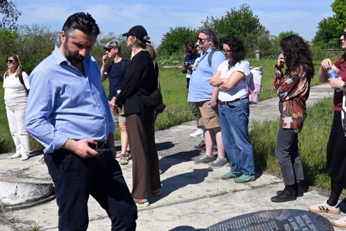 Group of adults standing in a line along a sunny outdoor path, some looking at their phones, with grass and trees in the background.