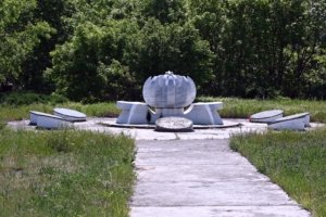 Abstract white sculpture with a segmented dome on a pedestal, set in a grassy, overgrown outdoor area with trees behind.