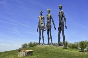 Four tall, weathered human statues stand on a grassy hill with a stone memorial labeled 1941–1945 in front.