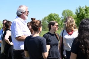 Group of people outdoors on a sunny day, standing together and chatting in a park-like setting with trees in the background.