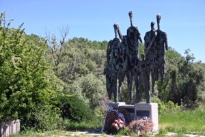 Group of four tall, gaunt statues in a memorial, with wreaths at the base and lush greenery around them.