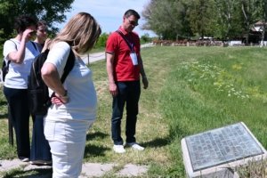 Group of adults outdoors examining a stone memorial plaque in a sunny grassy park with trees in the background