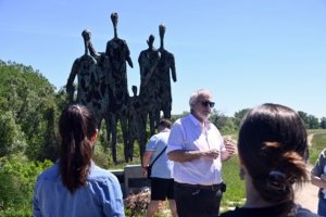 A group of people standing outdoors around a tall, dark sculptural figure against a clear blue sky.