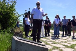 Group of adults outdoors on a sunny day; a man with white hair and beard speaks to the group from the foreground while others listen nearby.