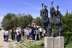 Group of people gathered in a sunny park, looking at a tall bronze sculpture of abstract standing figures.