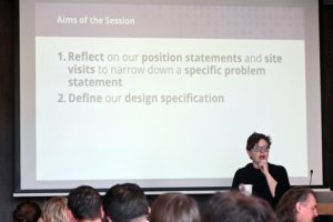 Presenter at a podium speaks to an audience; slide title reads 'Aims of the Session' with two bullet points visible.