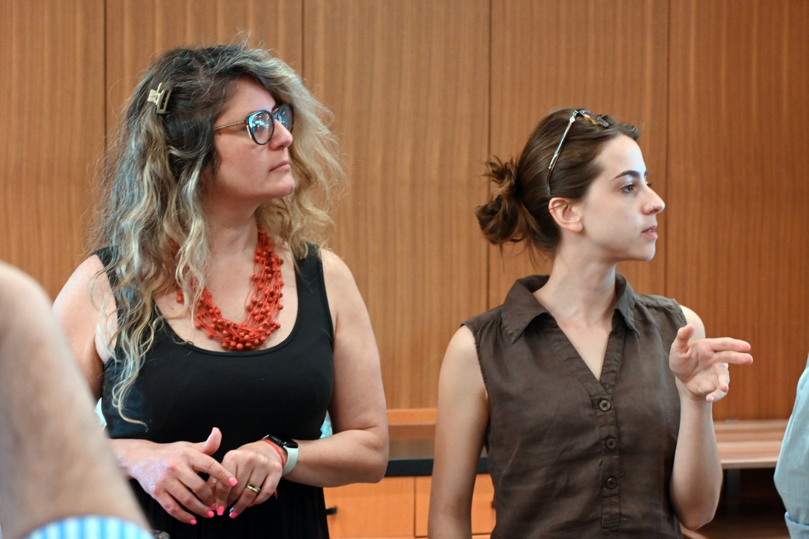 Two women stand side by side in a wood-paneled room; left woman wears a black dress with a red beaded necklace, right woman in a brown sleeveless shirt with sunglasses on her head.