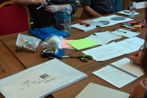 Cluttered conference table with papers, notebooks, markers, scissors, and a water bottle scattered across wood surface.