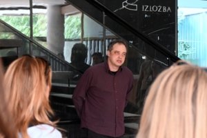 Man in a maroon shirt speaking to an audience in front of a black marble wall with the word IZLOZBA visible above the stairs.