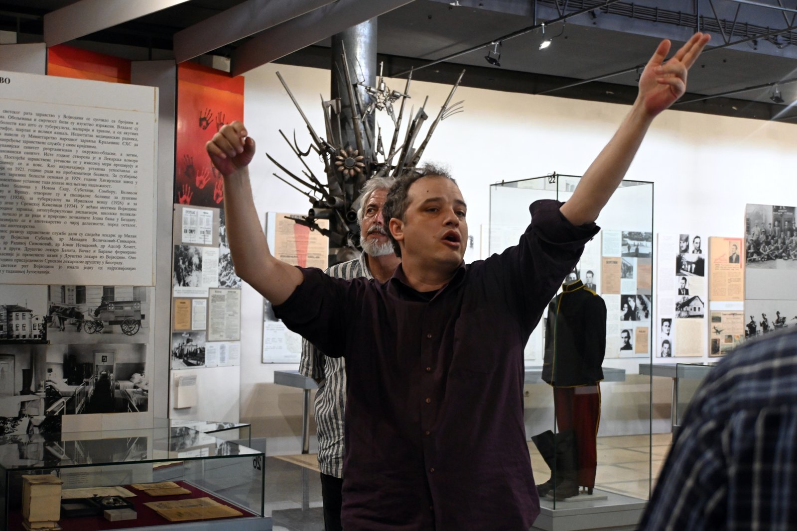 Man in a dark shirt raises both arms at a museum exhibit, with a spiky metal sculpture behind him and glass display cases around.