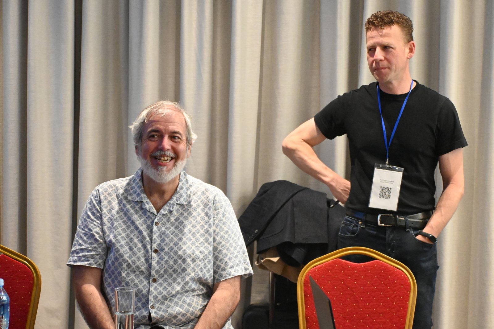 Older man with white beard and patterned shirt seated, smiling, as a younger man in a black t-shirt with a blue lanyard stands beside him at a conference backdrop of beige curtains.