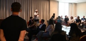 Speaker with grey hair addressing a conference room audience at the front of the room.