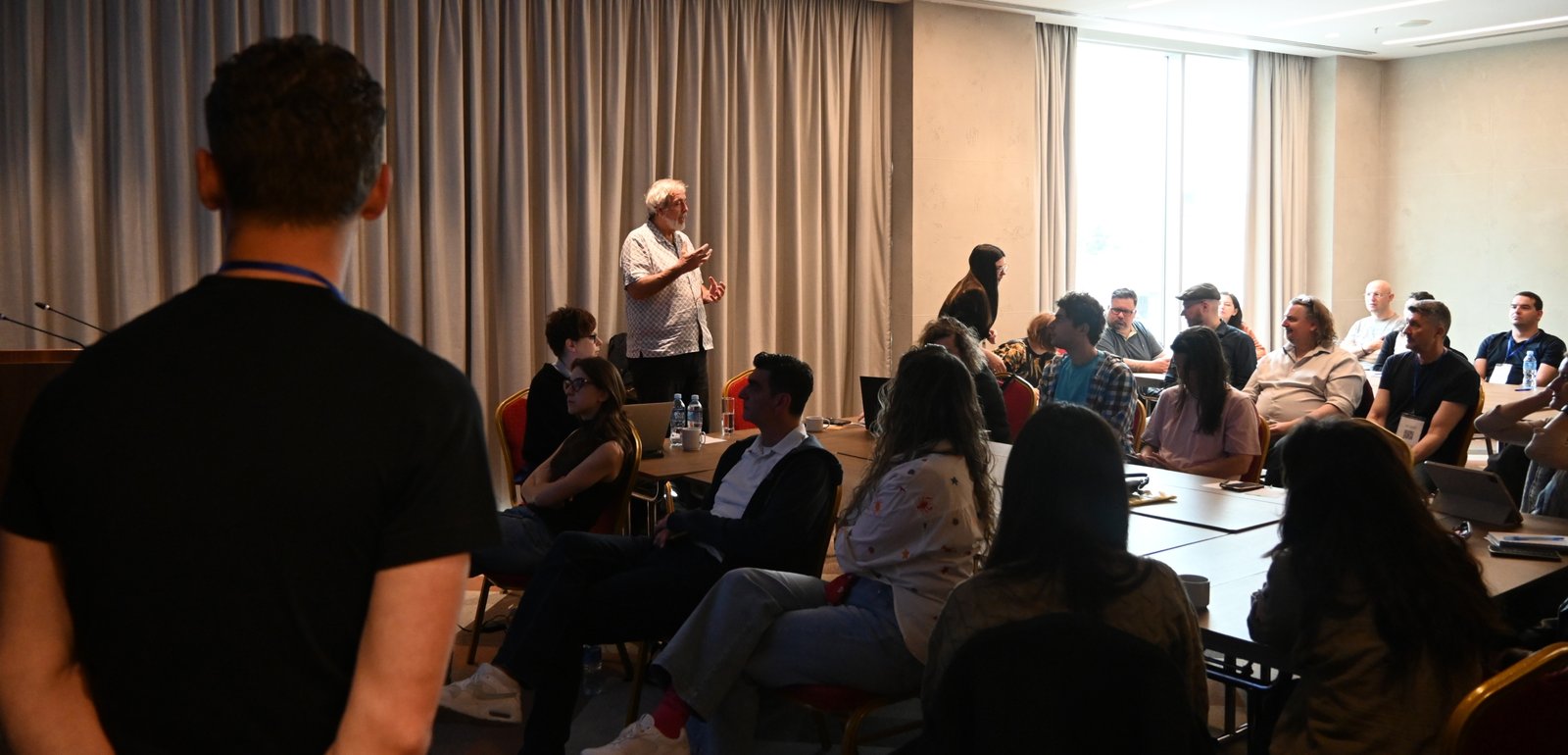 Speaker with grey hair addressing a conference room audience at the front of the room.