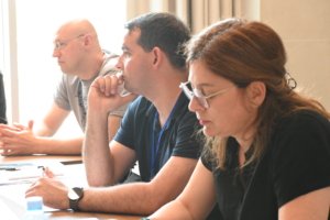 Three adults sit at a conference table in discussion, with a man in a navy shirt center and a woman with glasses in the foreground.