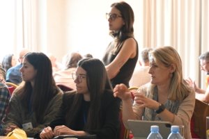Woman with glasses standing among seated attendees at a meeting or workshop, mid-conversation in a bright room with curtains behind.