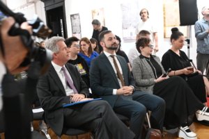 Audience seated in a bright room during a formal event, many in suits and smart casual wear with posters on the walls in the background.