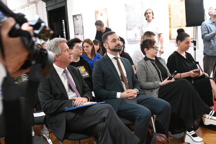Audience seated in a bright room during a formal event, many in suits and smart casual wear with posters on the walls in the background.