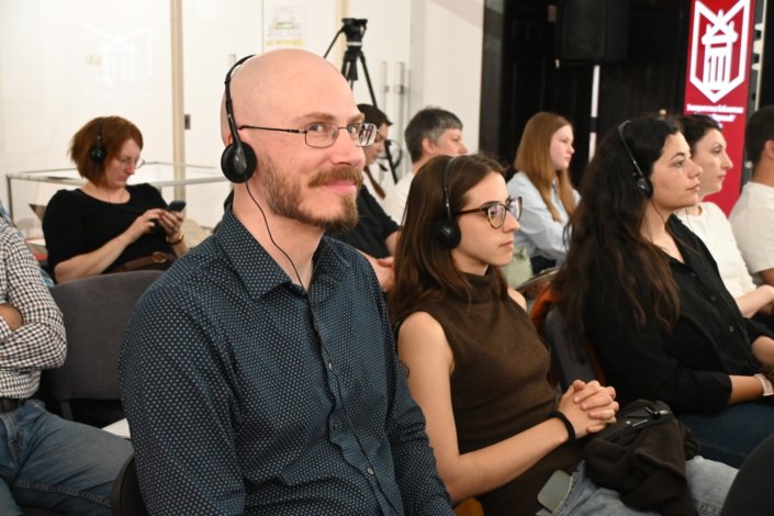 Bearded man with glasses wearing a headset in the foreground, others with headsets seated behind him in a conference setting.
