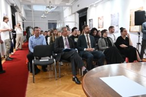 Audience seated in a bright gallery-like room, photographers at left capturing the event; a round wooden table in the foreground.