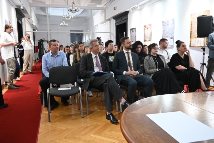 Audience seated in a bright gallery-like room, photographers at left capturing the event; a round wooden table in the foreground.