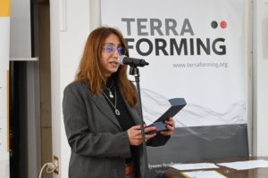 Woman with red hair and glasses speaking into a microphone at a conference, standing in front of a 'TERRA FORMING' banner.