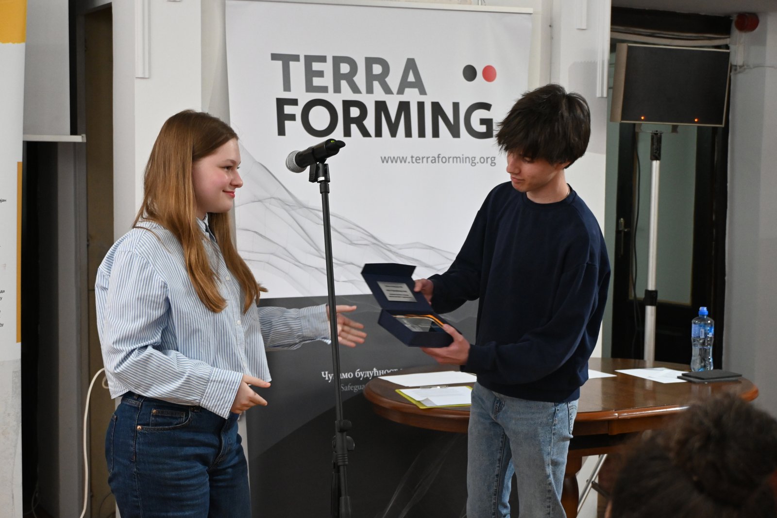 Two young people on stage exchanging a plaque or award in front of a Terra Forming banner with a microphone nearby.