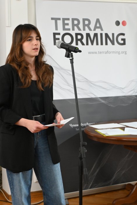 Young woman speaks into a microphone at a Terraforming event, with the banner reading 'TERRA FORMING' behind her.