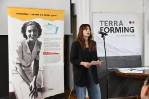 Woman speaking into a microphone at a presentation, with a large vintage photo banner on the left and a 'TERRA FORMING' banner on the right.