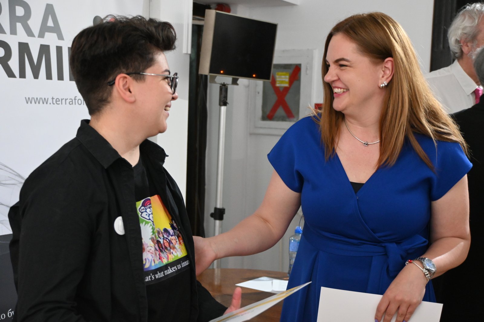 Two people smiling and shaking hands at a meet-and-greet event, papers in hand, indoors.