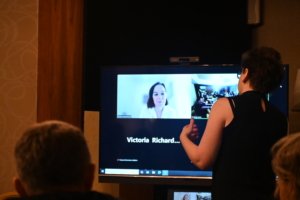 Person standing in front of a large monitor during a video conference, screen showing a woman speaking to the group.