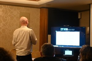 Bald man with glasses presenting to an audience in a conference room, standing before a large screen displaying slides.