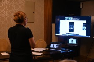 Back view of a presenter in a black shirt speaking into a microphone while a large monitor shows a slide deck on a desk setup in a conference room.