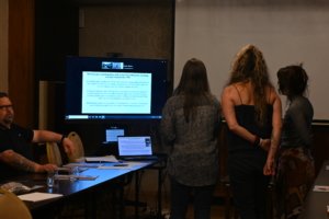 Group of people standing and watching a presentation on a large monitor in a conference room.