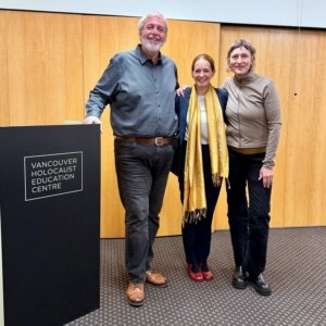 Three people stand together indoors, smiling for the camera, with a podium reading 'Vancouver Holocaust Education Centre' on the left.