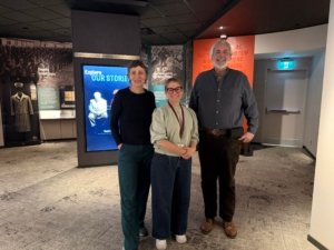 Three adults stand together in a museum exhibit space, smiling, with large historical display panels behind them and a touchscreen screen nearby.