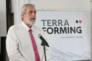 Bearded man in a white shirt and pink tie speaking into a microphone at a TerraForming event.