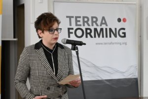 Woman with short brown hair and glasses speaks into a microphone at a TerraForming event, wearing a black-and-white checkered blazer.