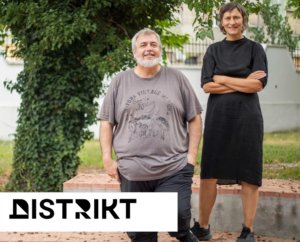 Two adults posing outdoors, a smiling man with a gray beard in a gray T‑shirt and a woman in a black dress, with a white Distrikt logo overlay in the corner.