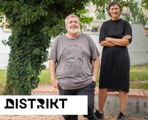 Two adults posing outdoors, a smiling man with a gray beard in a gray T‑shirt and a woman in a black dress, with a white Distrikt logo overlay in the corner.
